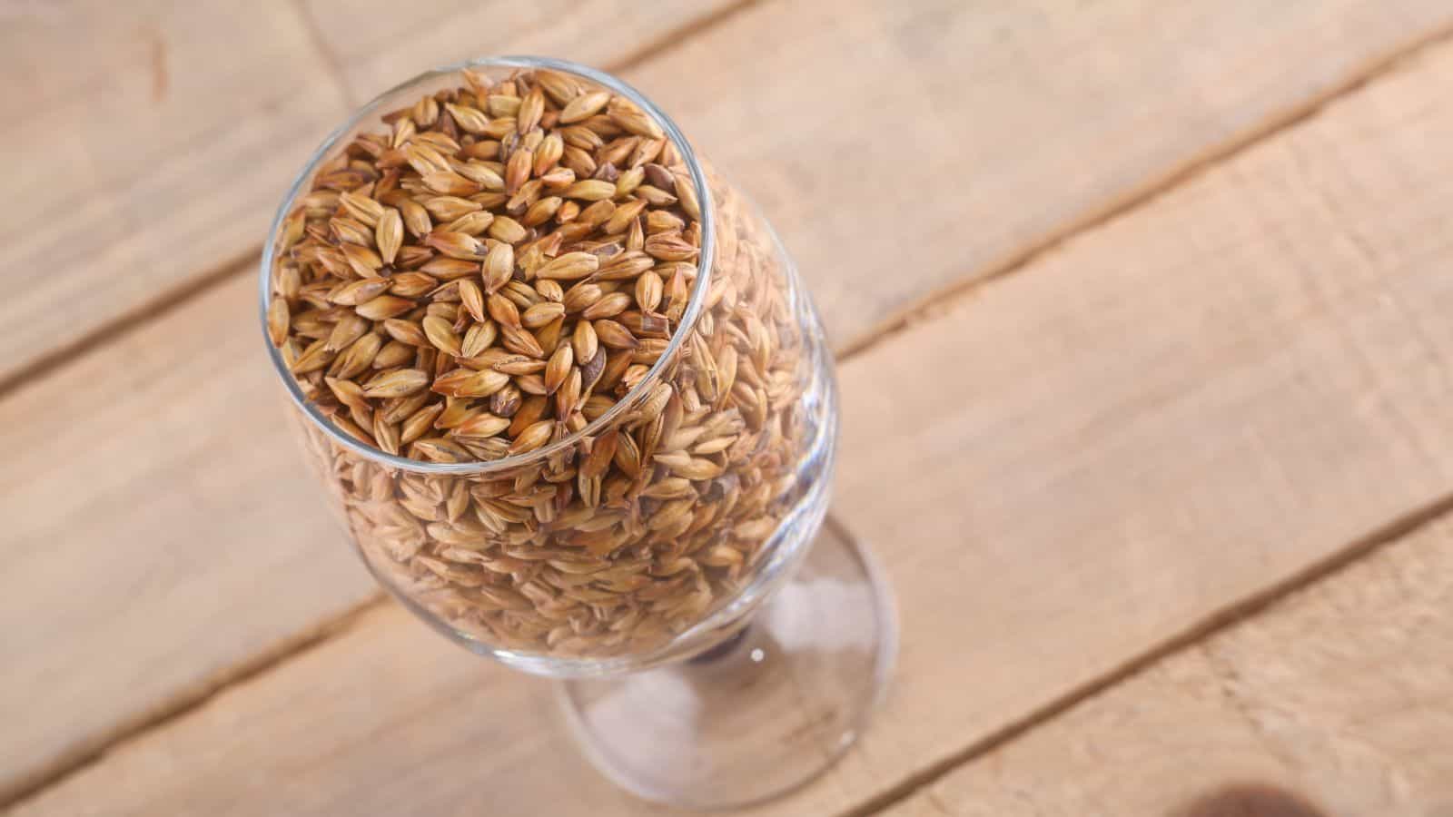 A transparent glass filled with barley grains is placed on a wooden surface. The image is taken from above, showing the texture and color of the grains inside the glass.