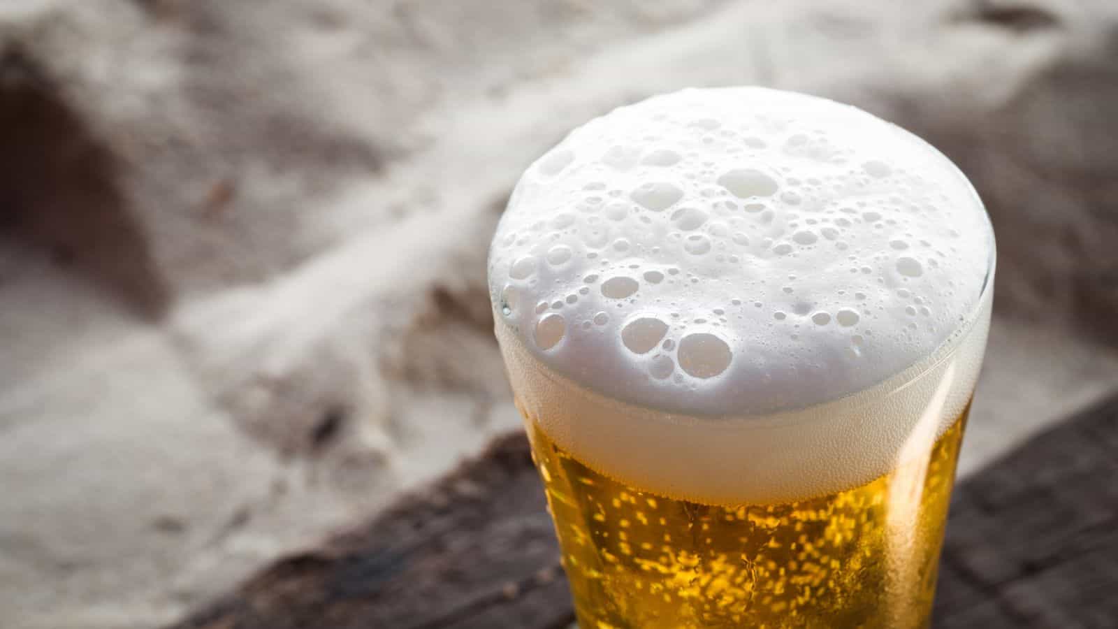 A glass of beer with a thick, frothy head sits on a wooden surface. The beer is golden and translucent. Soft-focus sand is visible in the background, suggesting an outdoor, beach setting.