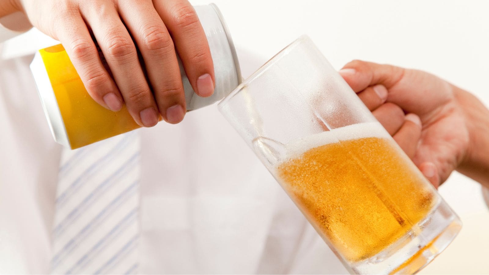 A person in a white shirt pours beer from a can into a clear glass. Both hands are visible, with one holding the can and the other holding the glass tilted to receive the beer. The background is plain and light-colored.