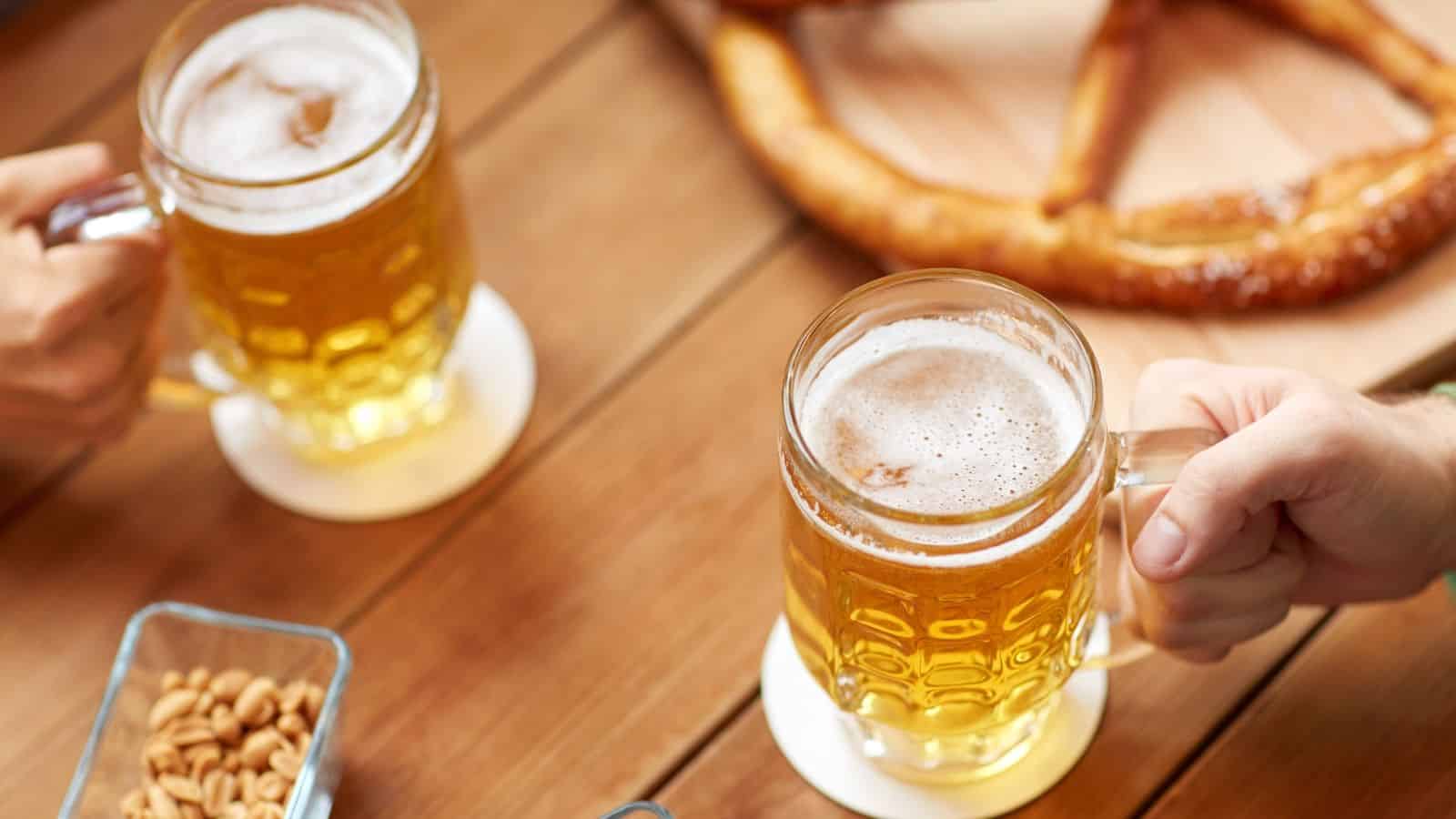 Two people are holding mugs of beer on a wooden table. There is a large pretzel and a small bowl of nuts in the background. The scene suggests a casual, social setting.