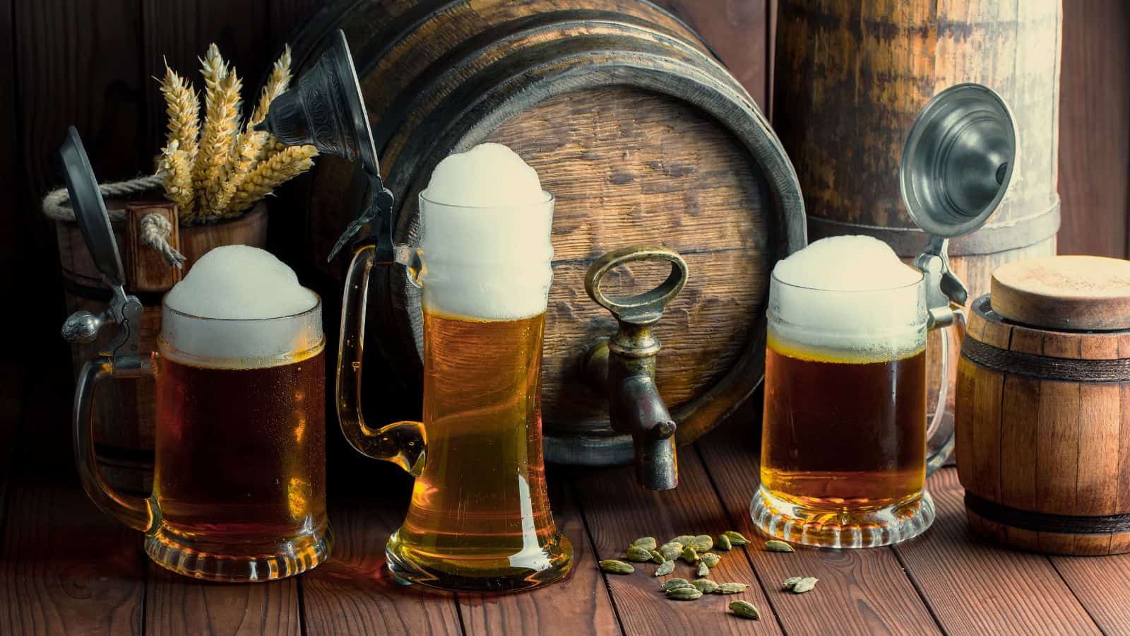 Three glass mugs filled with frothy beer are placed in front of wooden barrels. A bundle of wheat and loose hops are on the wooden surface, creating a rustic atmosphere. The open lids of the mugs are visible.