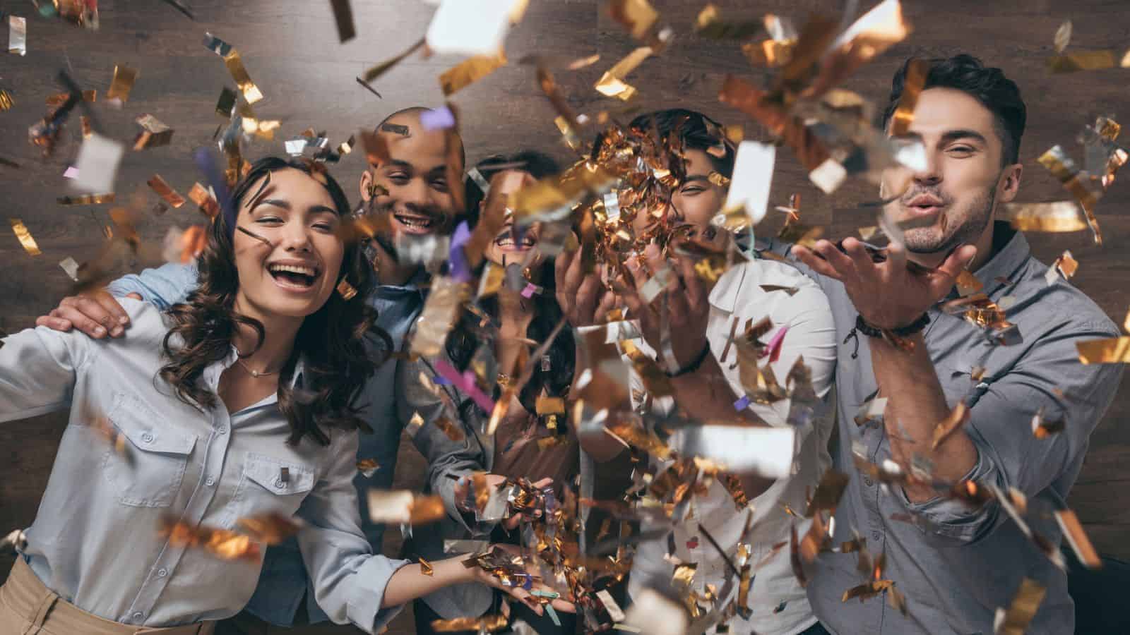 A group of five people smiling and celebrating while surrounded by falling confetti. They appear to be in a festive environment, creating a joyful atmosphere. The background is a wooden wall.