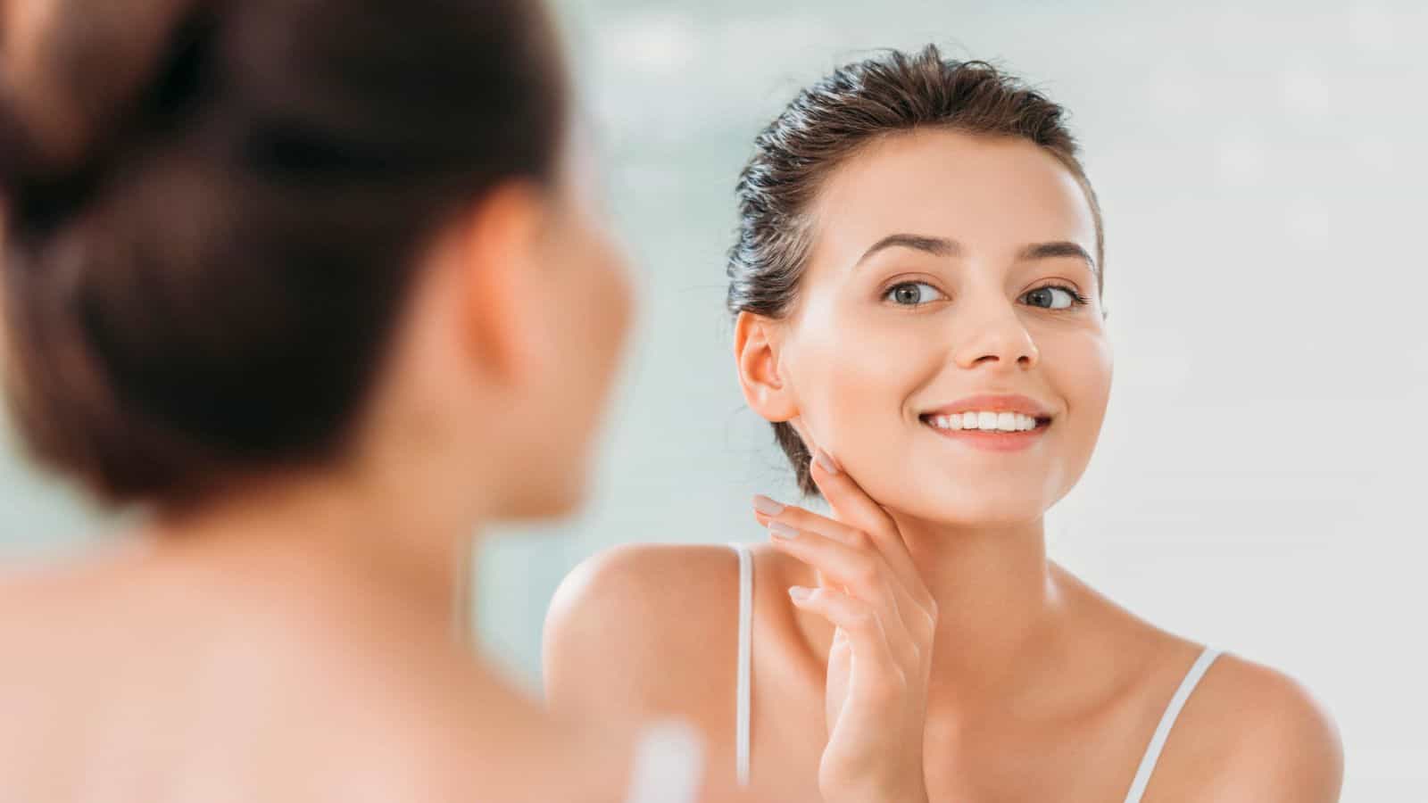 A woman smiles and touches her face while looking into a mirror. She has her hair tied back and is wearing a white tank top. The background is softly blurred, highlighting her reflection.