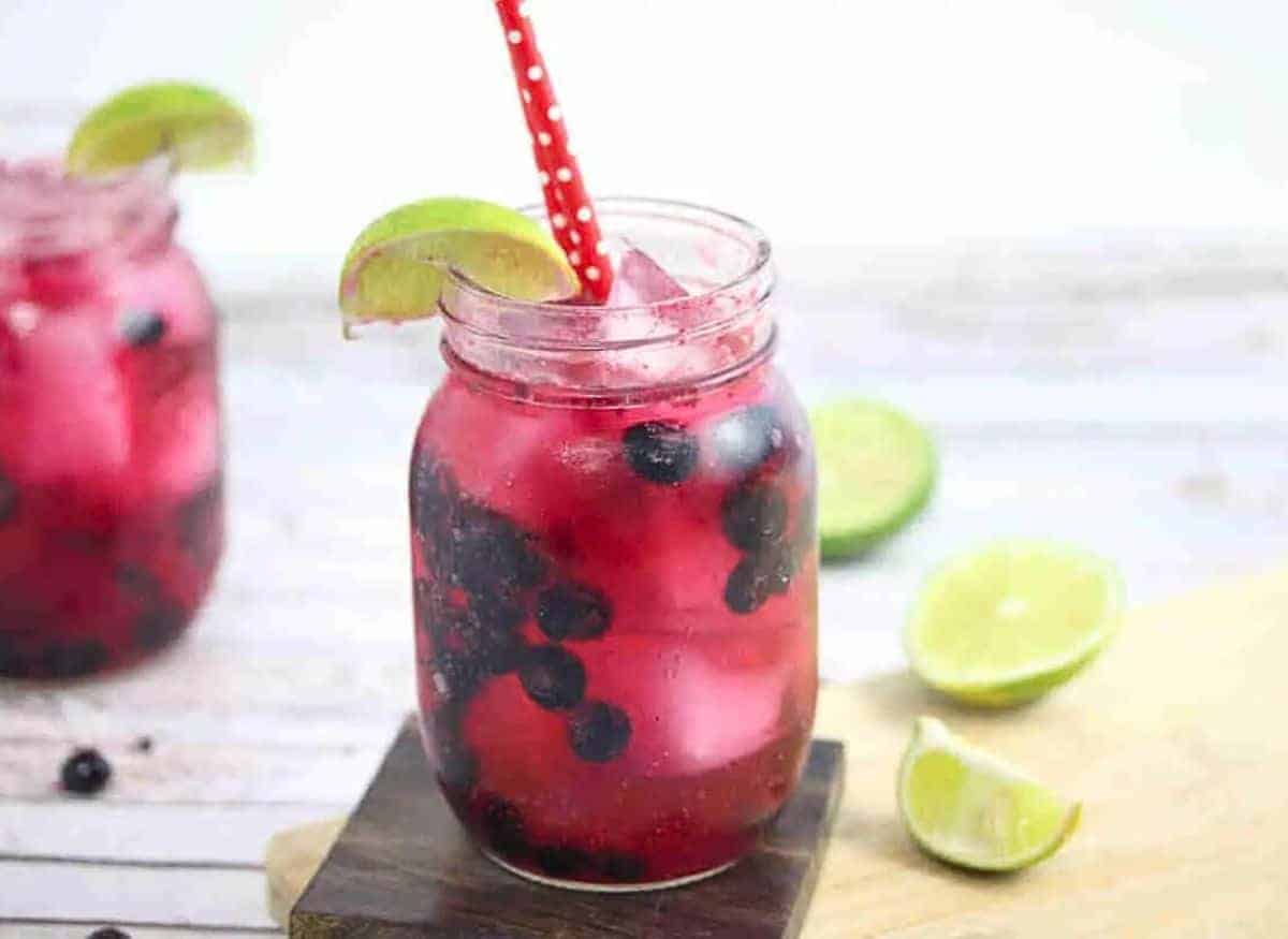 A mason jar filled with pink lemonade, ice cubes, and blueberries, garnished with a lime wedge and a red polka dot straw. Two lime wedges are on the wooden board beside it, with another jar in the background.
