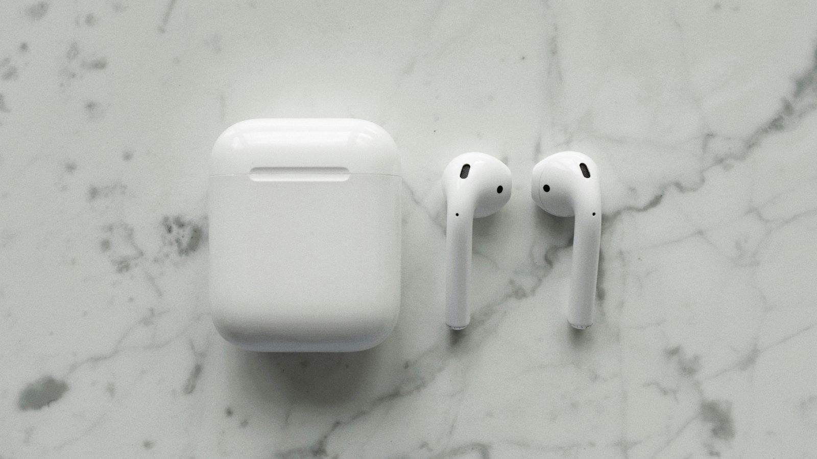A pair of white wireless earbuds next to their rectangular charging case is placed on a gray and white marble surface. The earbuds and case are minimalistic in design.