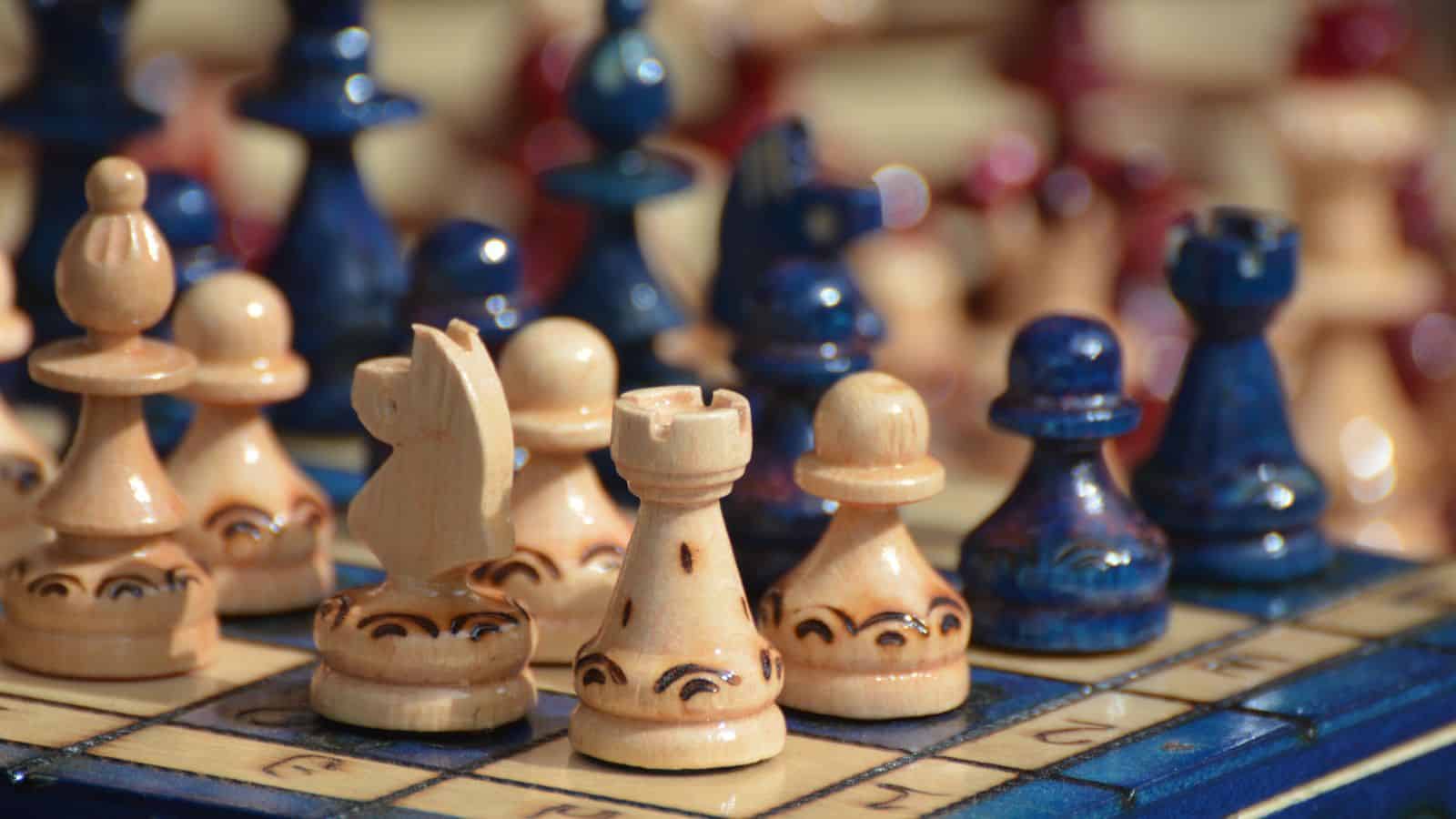Close-up of a decorative chess board with intricately carved wooden pieces. The pieces are painted in shades of cream and blue, set on a board with engraved letters and numbers. The focus is on the front row of pieces.