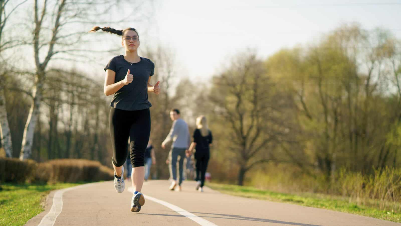 A person with a long braid is jogging on a paved pathway through a park. Two other people are walking in the opposite direction in the background. The trees lining the path have sparse leaves, indicating early spring.