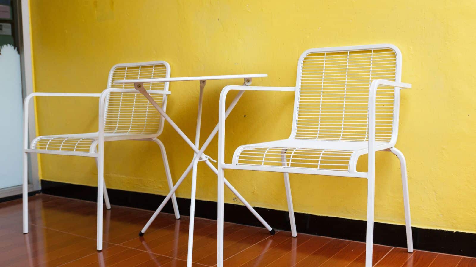 Two white metal chairs and a small white table are placed on a polished wooden floor against a bright yellow wall.