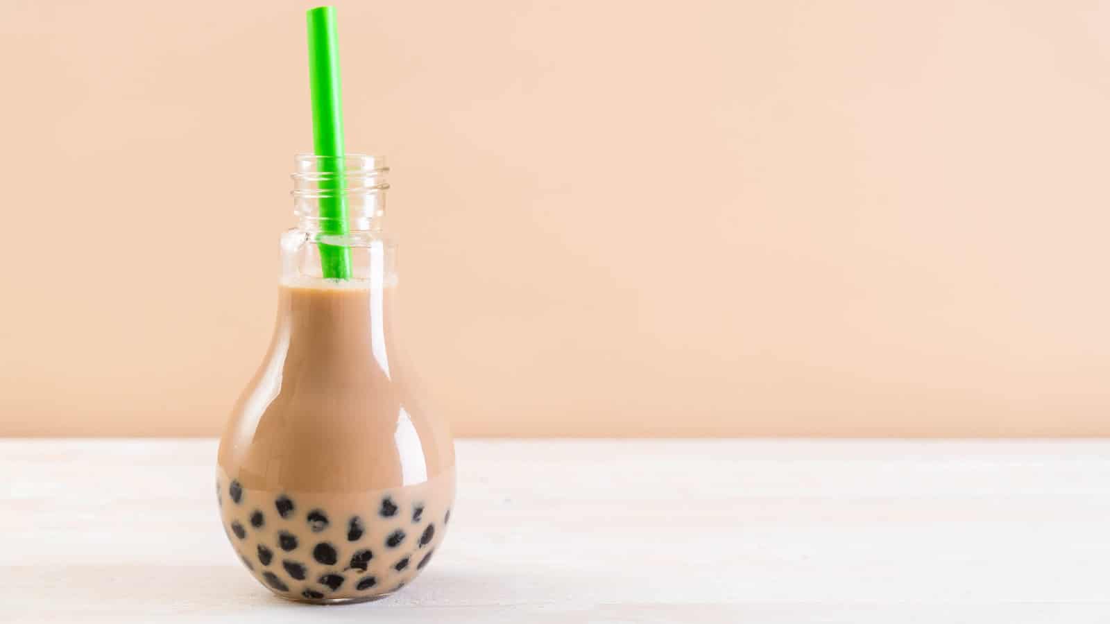 A light bulb-shaped glass filled with brown bubble tea and black tapioca pearls sits on a light wooden surface. A green straw is inserted into the drink. The background is a plain beige color.