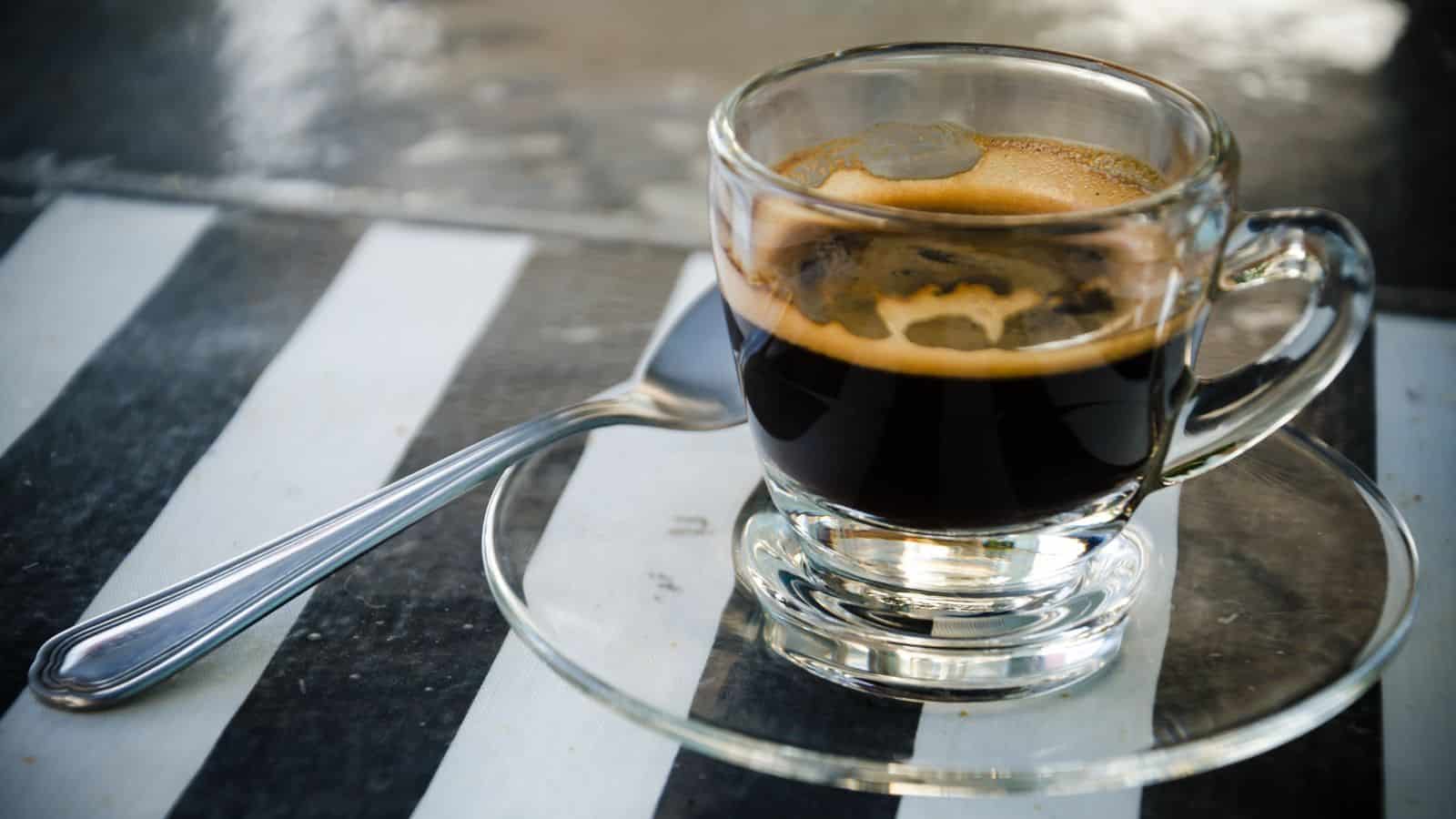 A small glass cup filled with espresso sits on a clear glass saucer. A silver spoon is placed beside the cup. The table underneath has a black and white striped pattern.