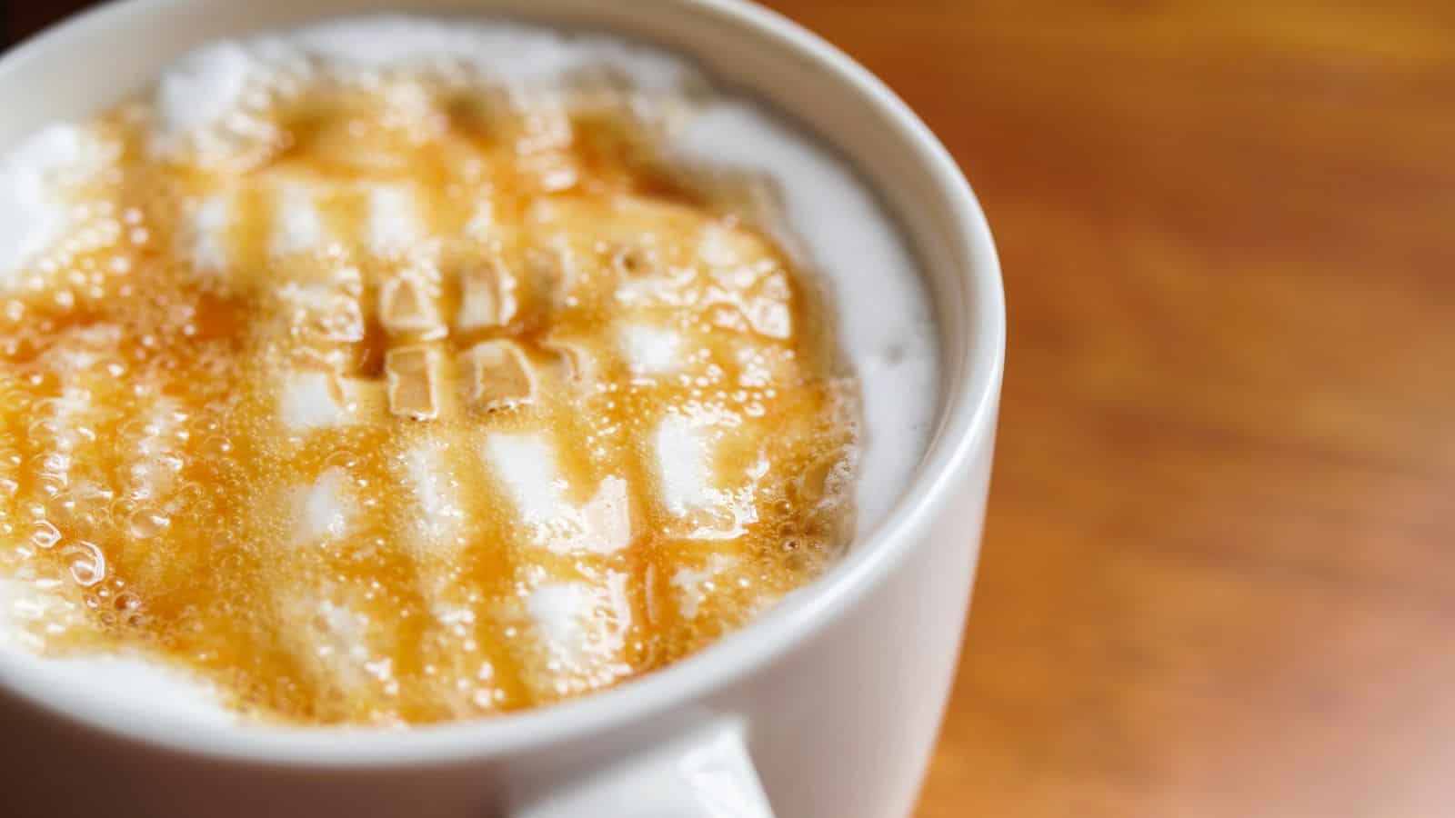 Close-up of a mug filled with a frothy coffee beverage topped with caramel drizzle, creating a lattice pattern. The mug is on a wooden surface with a warm, brown tone.