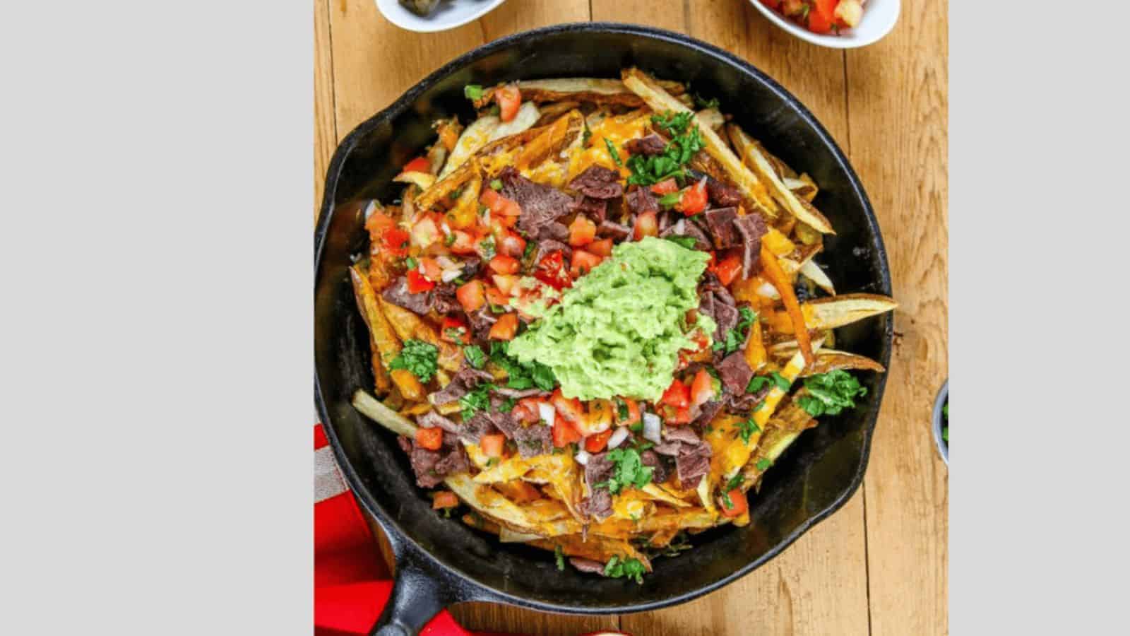 A cast-iron skillet filled with loaded fries, topped with melted cheese, diced tomatoes, blue corn chips, and a large dollop of guacamole. A red and white cloth is partially visible on the left, with small bowls in the background.