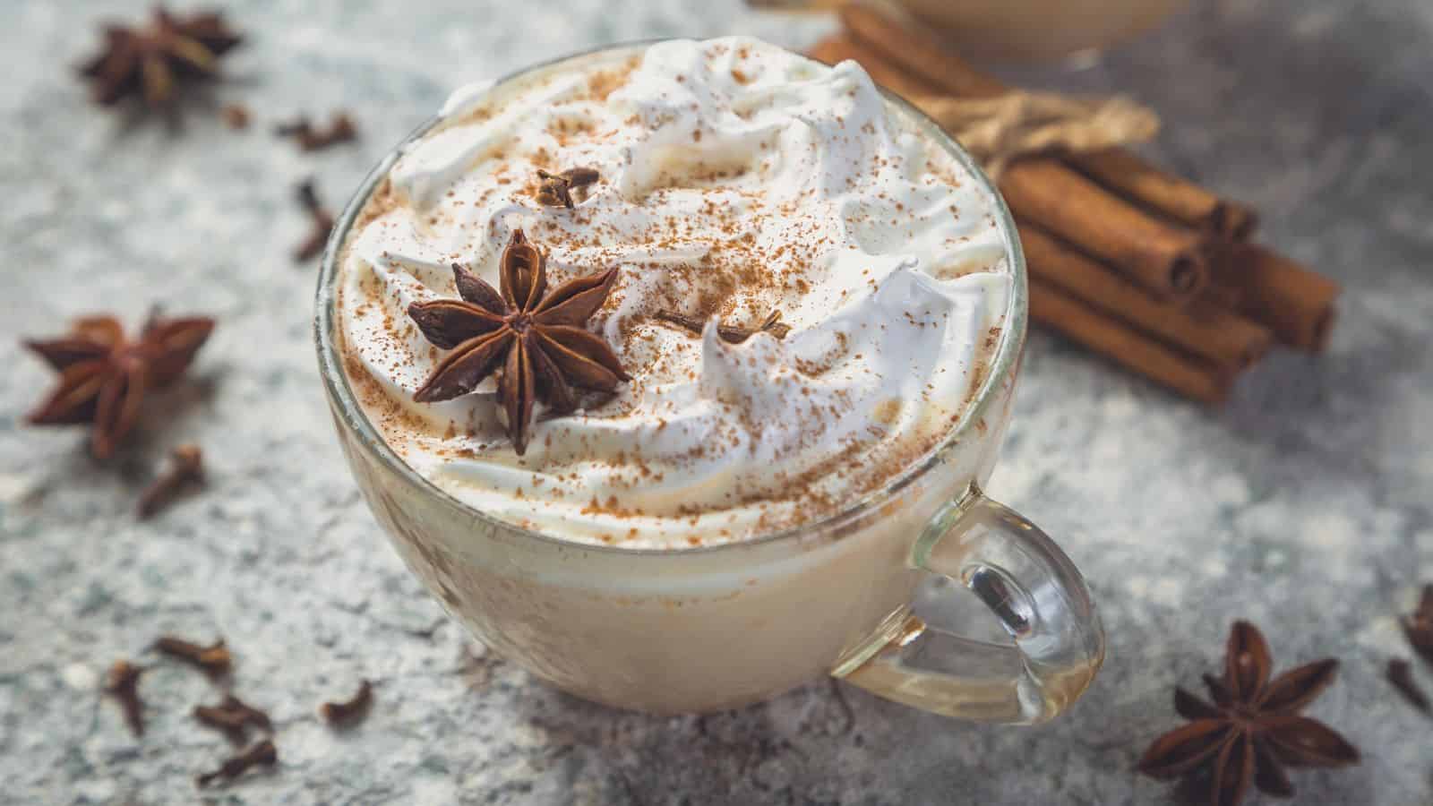 A glass mug filled with a creamy beverage topped with whipped cream and sprinkled with cinnamon. A star anise sits atop the cream. Cinnamon sticks and additional star anise are scattered on the textured surface nearby.