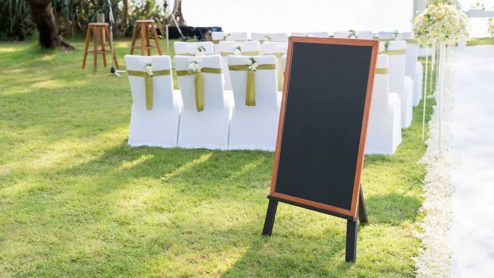 An outdoor ceremony setup on grass features a blank blackboard with a wooden frame in the foreground. Rows of white chairs with gold sashes are arranged, leading to an altar adorned with flowers in the background.