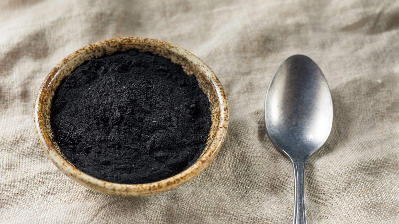 A ceramic bowl filled with black activated charcoal powder is placed on a beige fabric surface. A silver spoon is positioned to the right of the bowl.