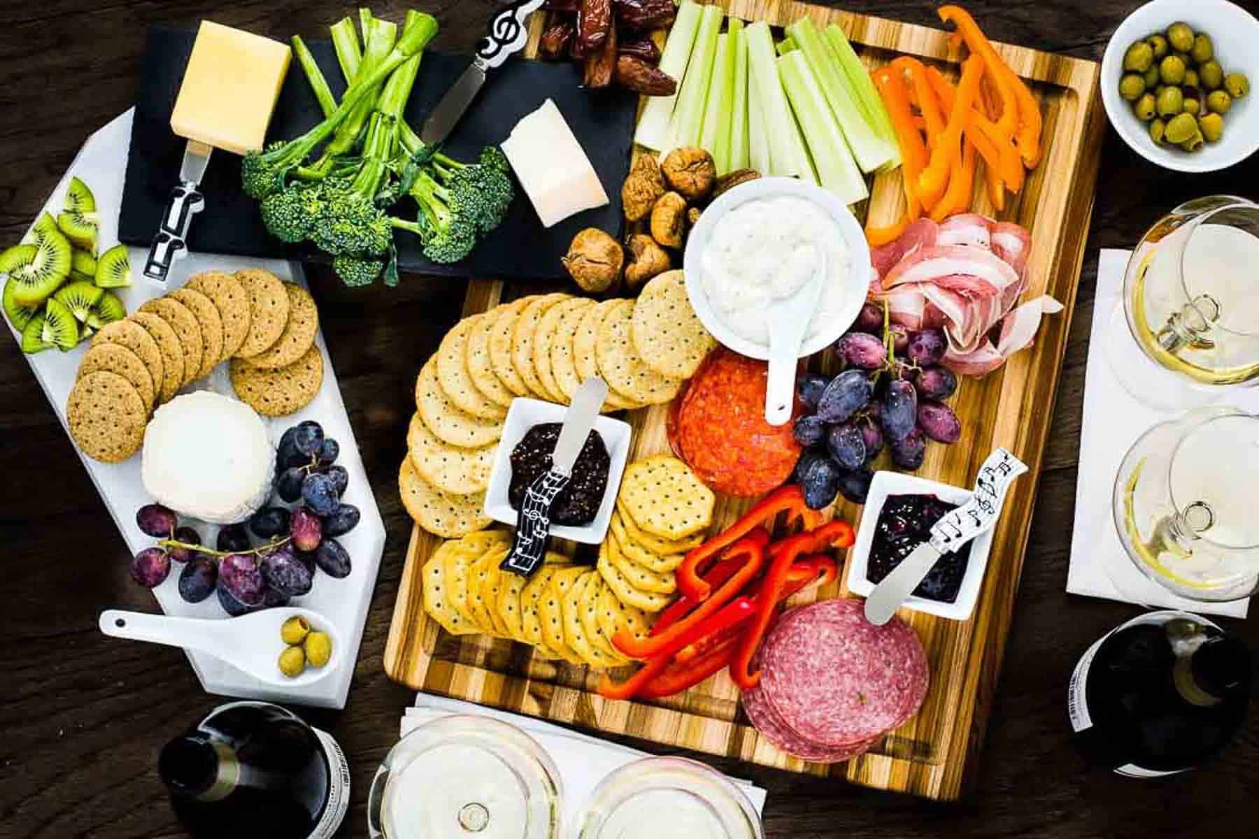 A variety of appetizers are arranged on a wooden board and white plates, including crackers, sliced meats, cheeses, vegetables, fruits, olives, and dips. Two glasses of white wine and a dark bottle are nearby on a dark table surface.