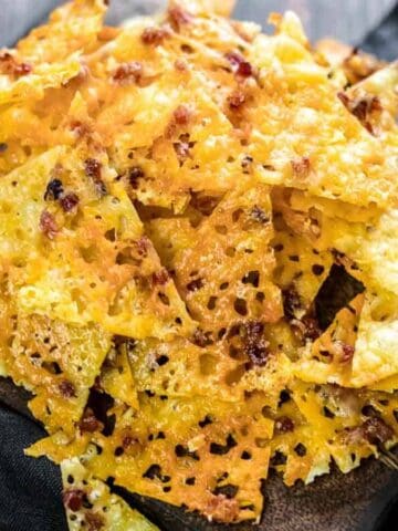 A pile of crispy, golden cheese crisps with sprinkled herbs and spices, arranged on a wooden board over a dark cloth. The crisps have a textured surface with small air bubbles. The background is a wooden table.