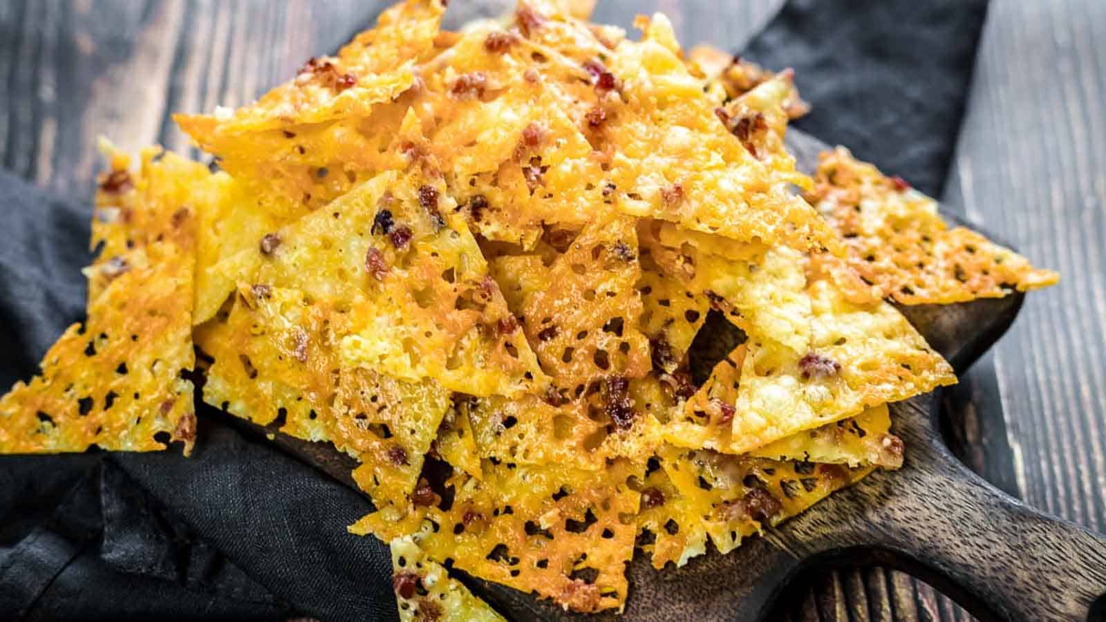 A pile of crispy, golden cheese crisps with sprinkled herbs and spices, arranged on a wooden board over a dark cloth. The crisps have a textured surface with small air bubbles. The background is a wooden table.