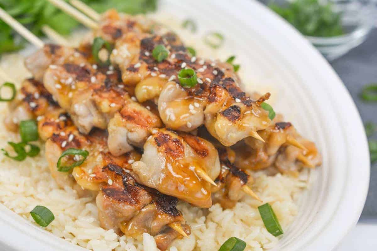 Grilled chicken skewers glazed with a shiny sauce are placed on a bed of rice. They are garnished with sliced green onions and sesame seeds, presented on a white plate.