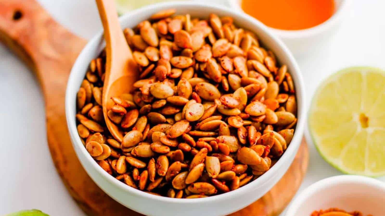 A bowl of seasoned, roasted pumpkin seeds with a wooden spoon on a wooden board. A half lime and a small dish of orange sauce are visible nearby.