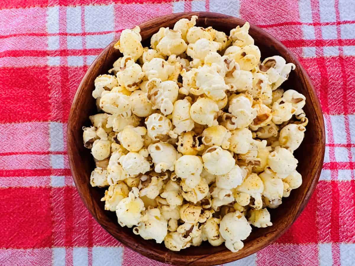 A wooden bowl filled with popped popcorn sits on a red and white plaid tablecloth. The popcorn appears seasoned with visible specks.
