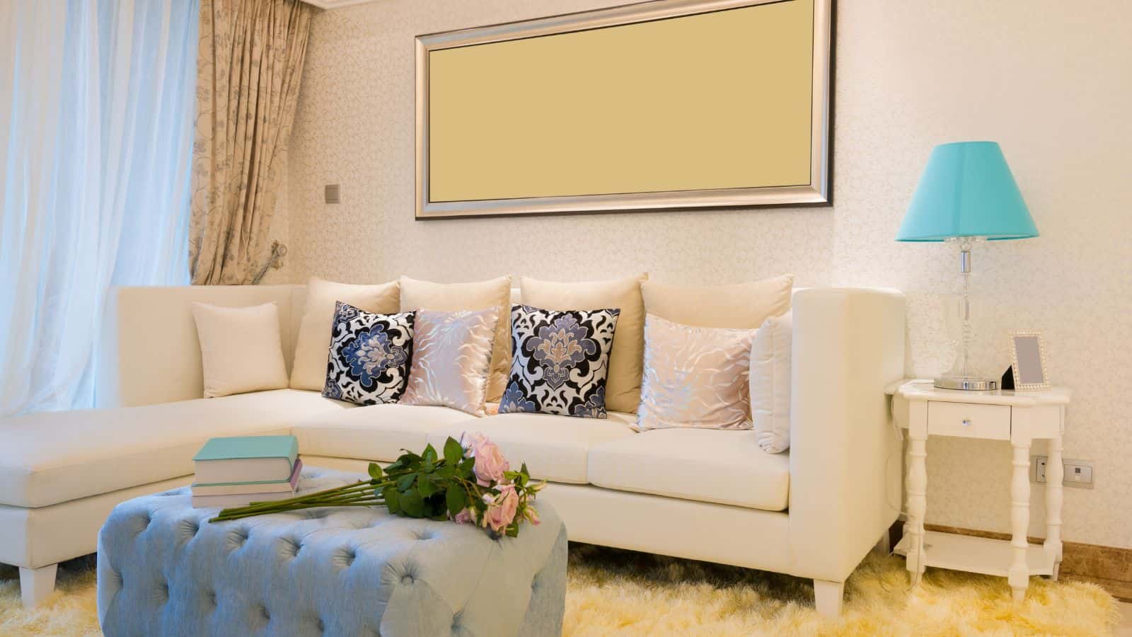 A living room with a white sectional sofa adorned with decorative pillows. A large empty picture frame hangs above. A light blue ottoman with a bouquet of flowers and books is in front. A side table with a blue lamp is on the right. Pale curtains cover a window.