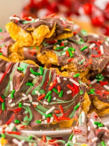 A plate of festive cornflake clusters topped with melted chocolate and multicolored sprinkles in red, white, and green. The background features a holiday-themed tablecloth and decorative red and green spheres.