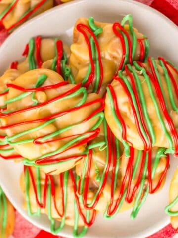 A plate of white chocolate-covered treats drizzled with red and green icing sits on a red and white checkered cloth. A glass of milk is visible nearby, and colorful spherical decorations surround the arrangement.