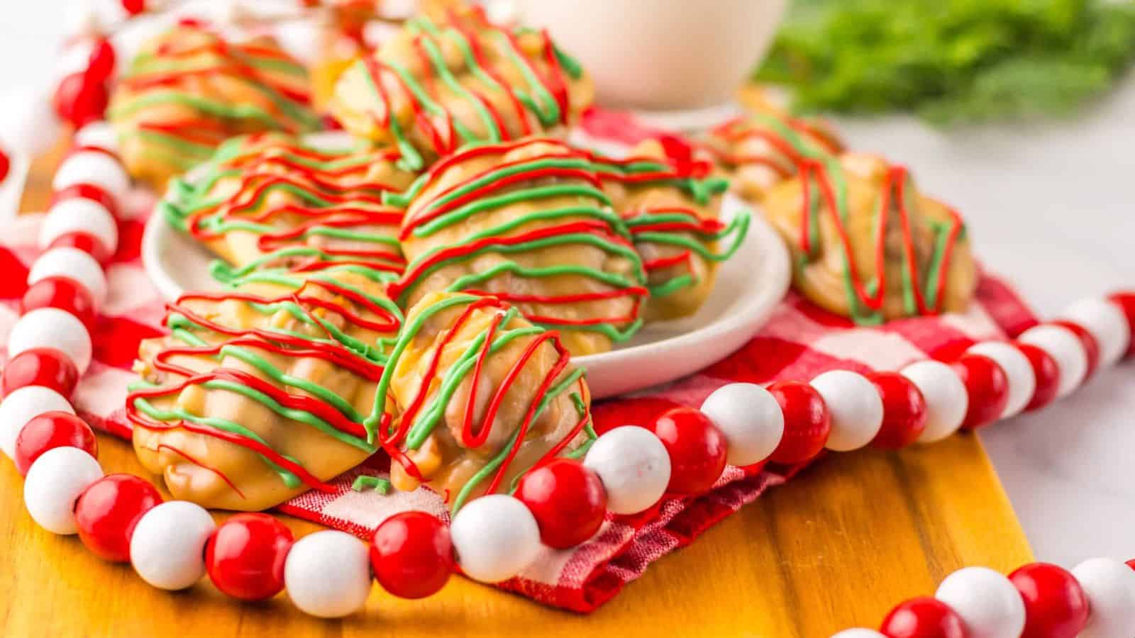 A plate of white chocolate-covered treats drizzled with red and green icing sits on a red and white checkered cloth. A glass of milk is visible nearby, and colorful spherical decorations surround the arrangement.