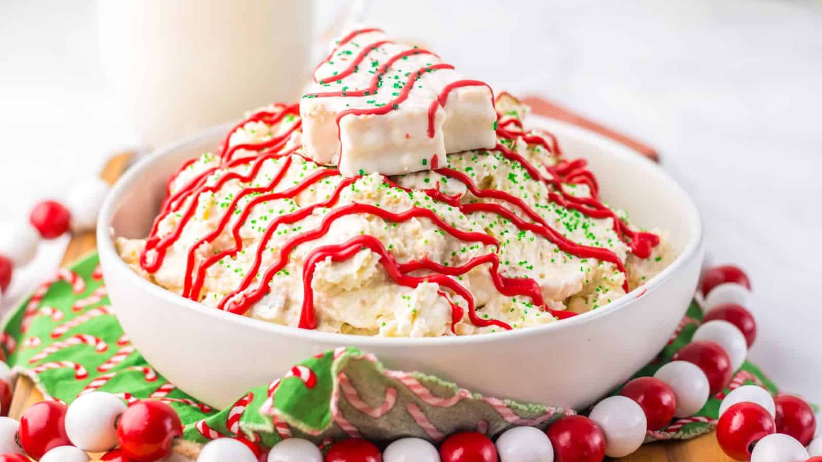 A bowl of holiday dessert featuring a creamy mixture topped with red and green sprinkles. A white chocolate-covered treat rests on top, drizzled with red icing. Candy beads and a festive napkin are in the background. Glass of milk is partially visible.