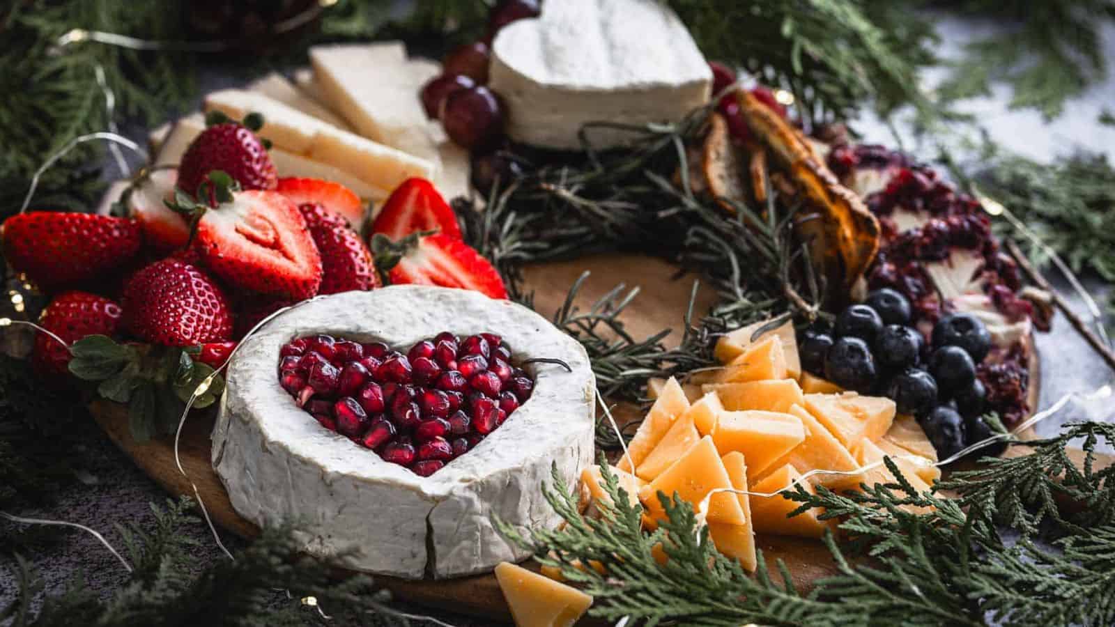 A cheese board featuring a wheel of brie with a heart-shaped cutout filled with pomegranate seeds. Surrounded by strawberries, grapes, blueberries, cheese slices, and decorative greenery.
