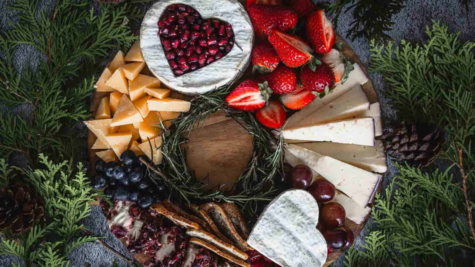 A charcuterie board with heart-shaped cheese topped with pomegranate seeds, surrounded by sliced strawberries, blueberries, grapes, assorted cheeses, crackers, and pine boughs.