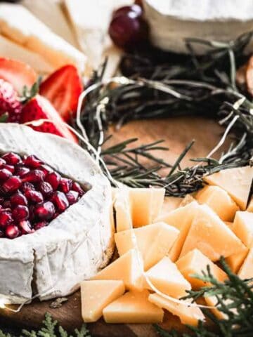 A cheese board with various cheeses, including a brie topped with pomegranate seeds, alongside strawberries, sliced cheese, blueberries, crackers, and a green garnish, presented on a wooden platter.