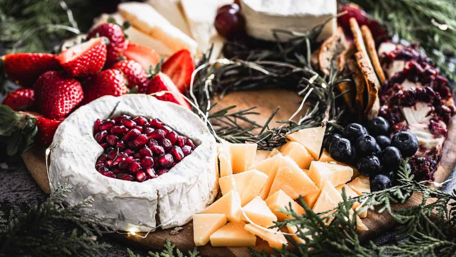 A cheese board with various cheeses, including a brie topped with pomegranate seeds, alongside strawberries, sliced cheese, blueberries, crackers, and a green garnish, presented on a wooden platter.