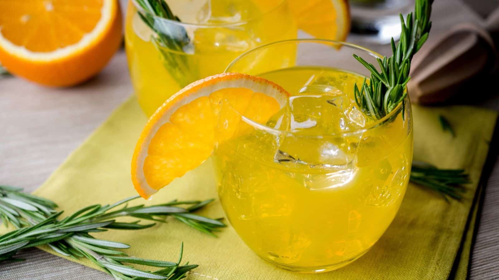 A glass filled with a bright orange drink, garnished with ice cubes, a slice of orange, and a sprig of rosemary. The drink is placed on a yellow napkin, with additional rosemary sprigs and an orange half visible nearby.