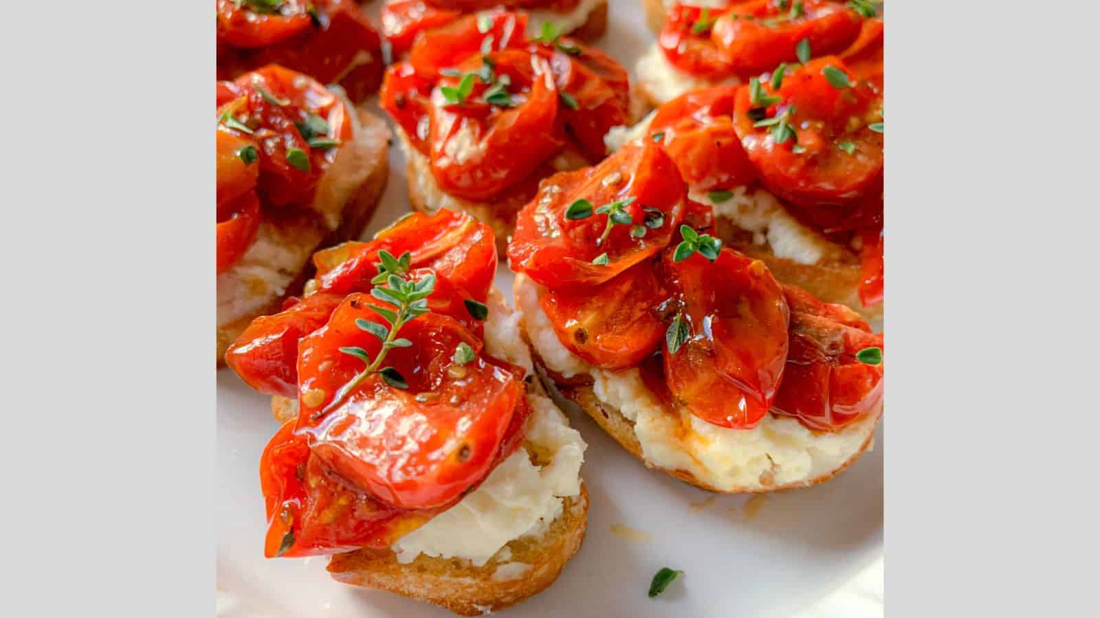 Slices of bread topped with creamy cheese, roasted cherry tomatoes, and fresh herbs are arranged on a plate. The tomatoes are glistening, contrasting with the smooth texture of the cheese and the crispy bread base.