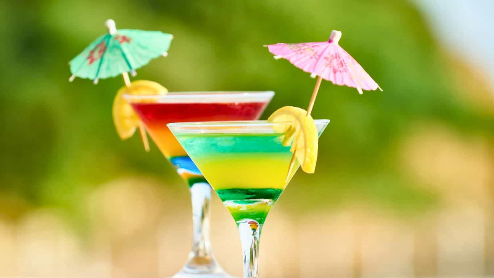 Two colorful cocktails in martini glasses with layered hues of red, yellow, green, and blue. Each drink is garnished with a small paper umbrella and a lemon slice. The background is blurred green, suggesting an outdoor setting.