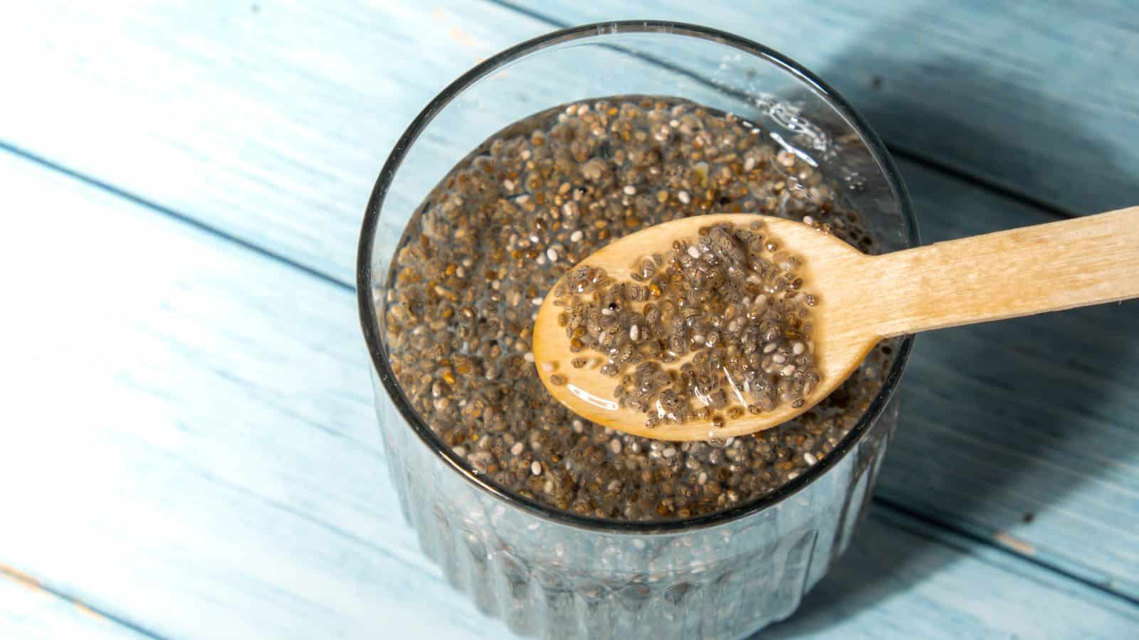 A glass filled with chia seeds soaked in water sits on a light blue wooden surface. A wooden spoon rests on the glass, holding soaked chia seeds. The chia seeds are swollen and have a gelatinous texture.