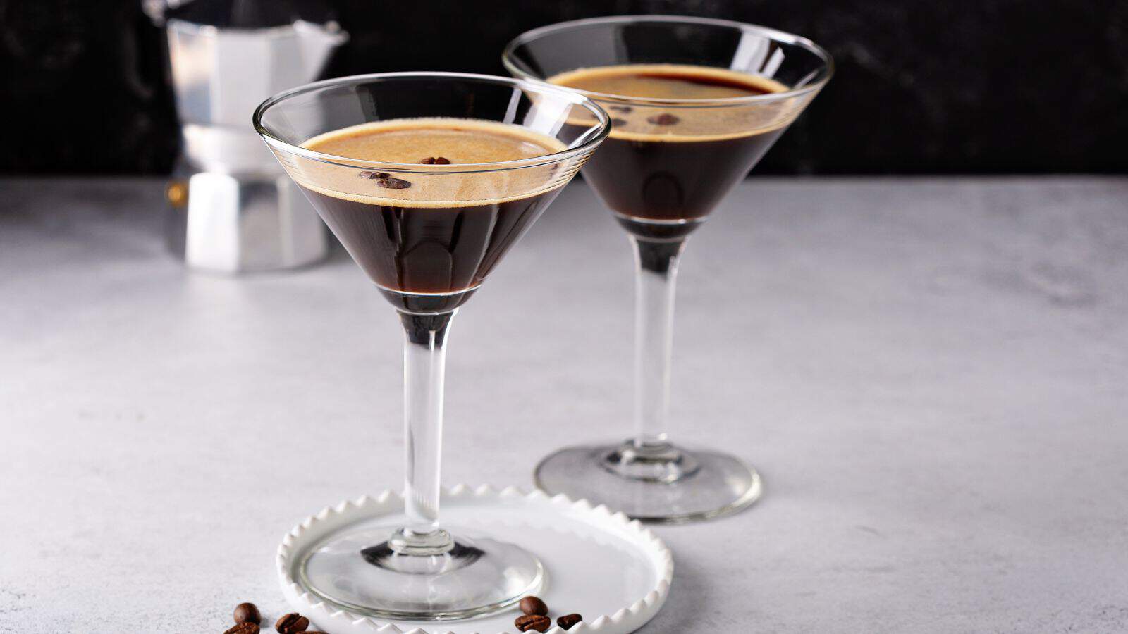 Two espresso martinis are served in cocktail glasses on a light gray surface. A small plate with coffee beans is placed in front of them, and a metallic espresso maker is blurred in the background.