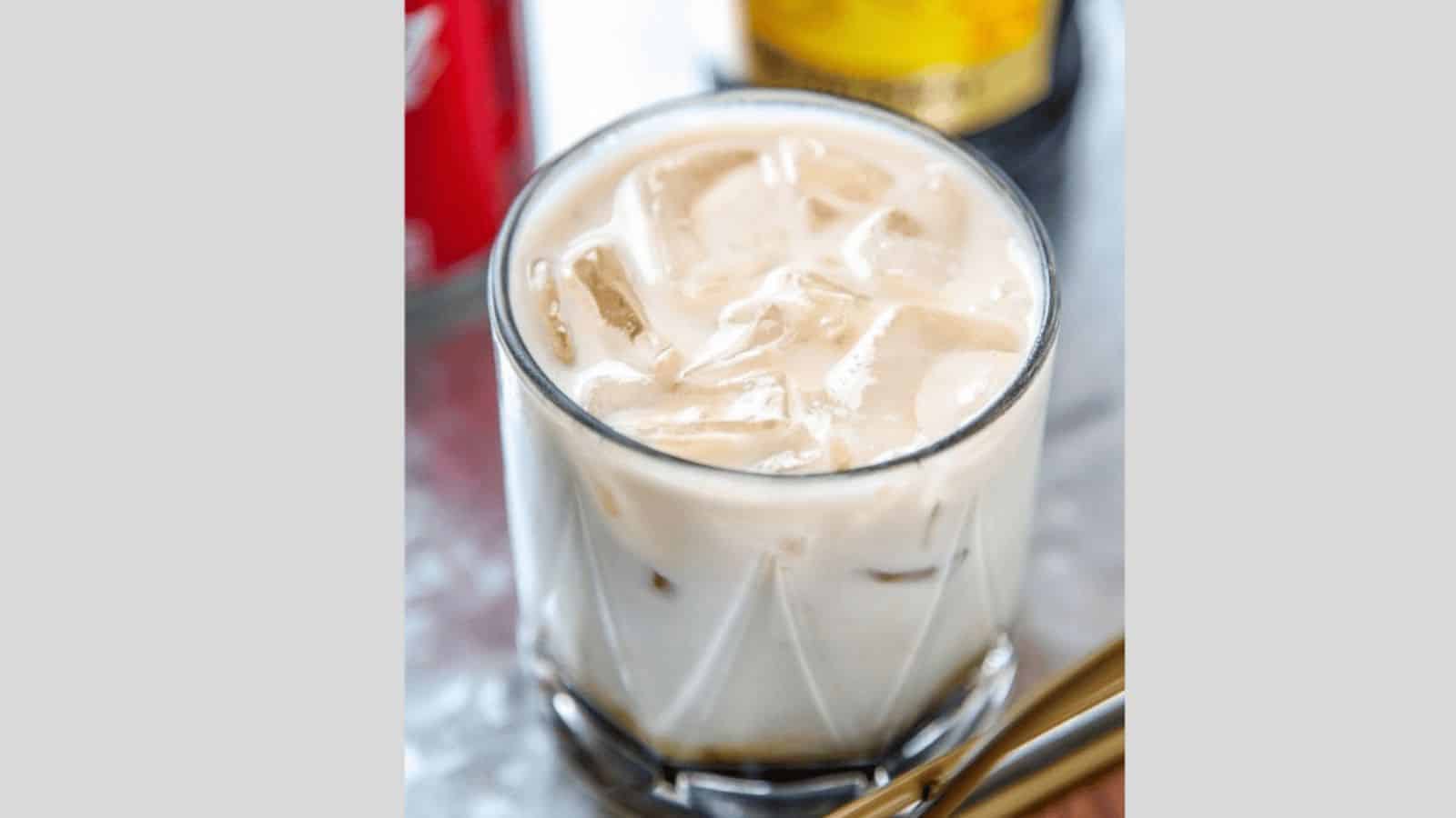 A glass filled with a creamy, iced beverage, likely a cocktail, sits on a table. The drink contains ice cubes and has a light tan color. A blurred background shows parts of a red can and a yellow-labeled bottle.