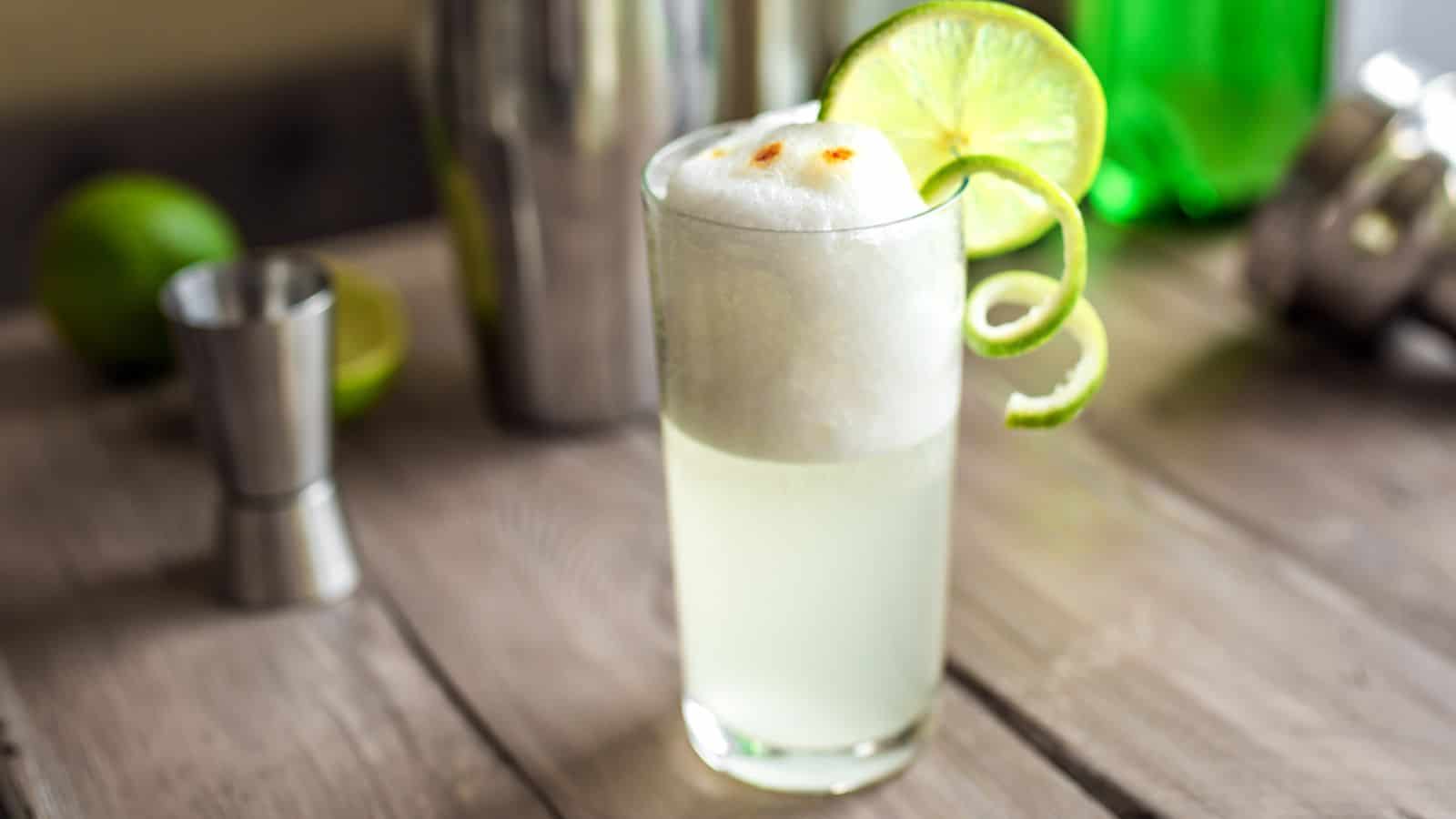 A tall glass of a foamy cocktail garnished with a lime wheel and a lime twist sits on a wooden table. A metal shaker, a jigger, and another lime are blurred in the background.