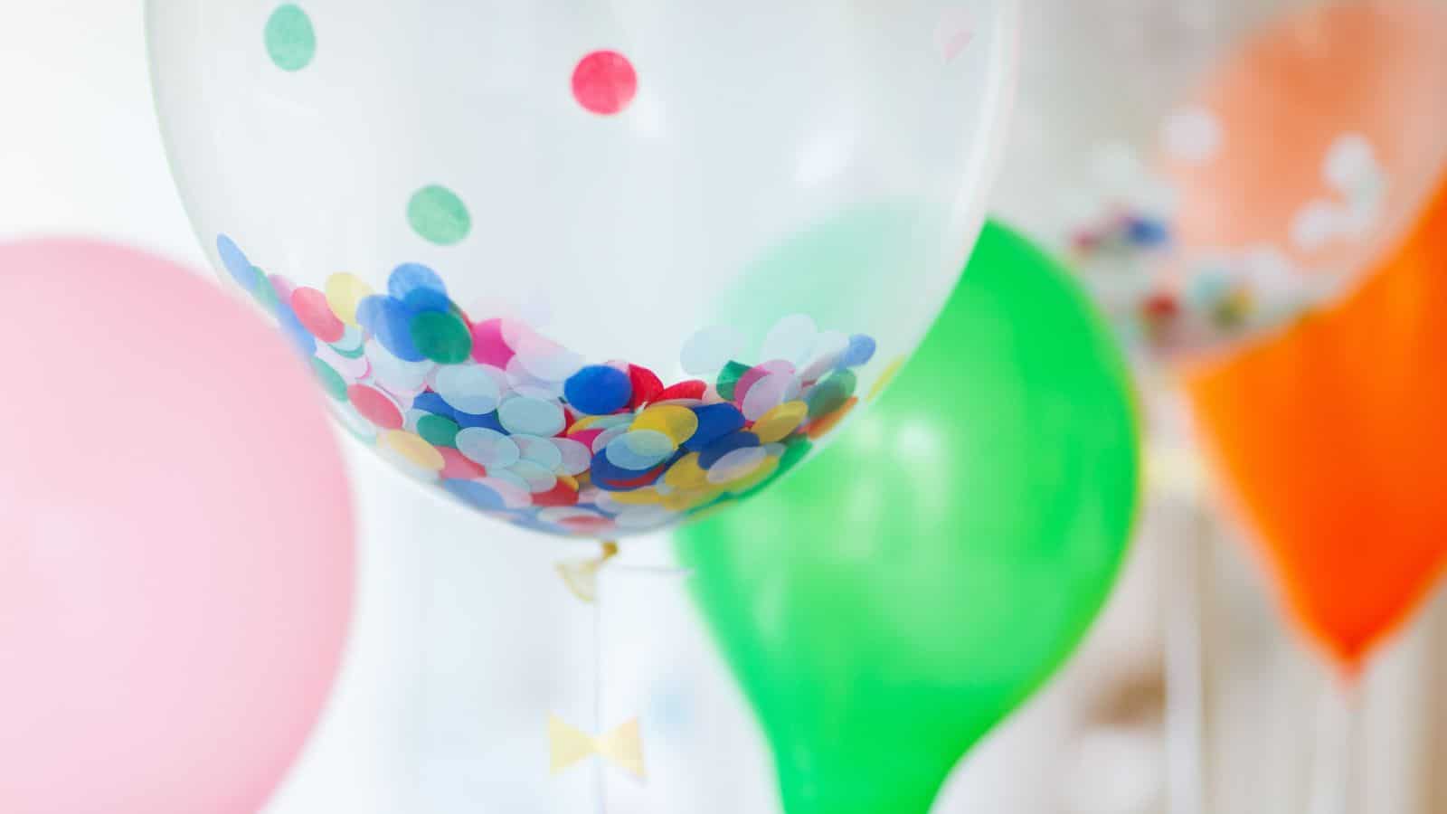 A close-up of a transparent balloon filled with multicolored confetti. In the background, there are additional balloons in pink, green, and orange. The scene is festive and colorful.