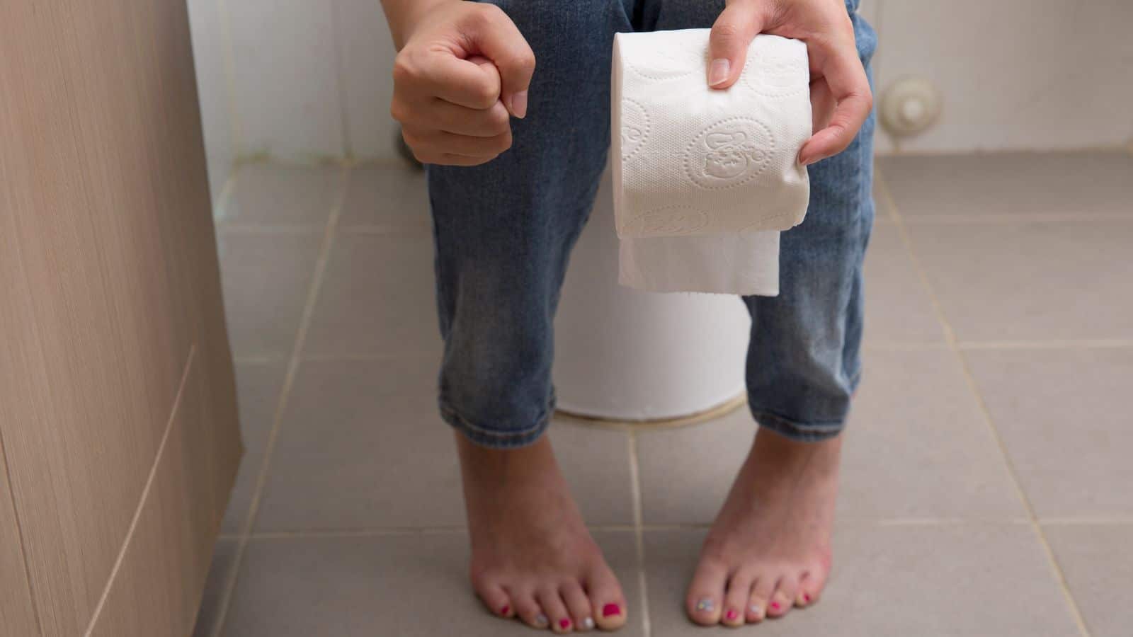 A person sitting on a toilet, holding a roll of toilet paper. They are wearing jeans and the floor is tiled. The person's feet are visible, and they have painted toenails in various colors.