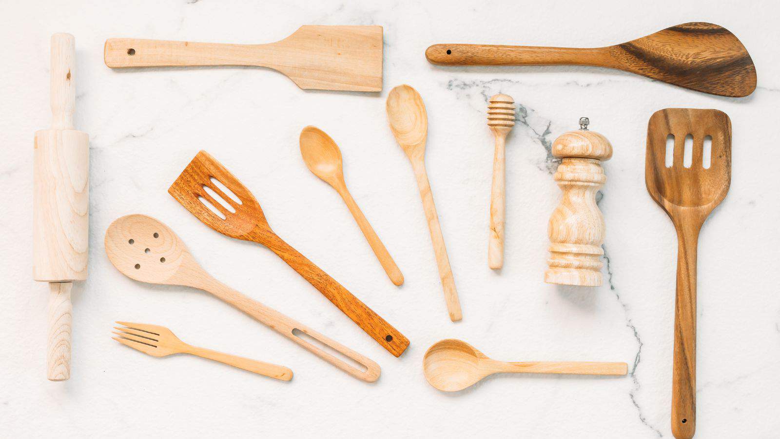 A collection of wooden kitchen utensils is arranged on a marble surface. Items include a rolling pin, spatulas, spoons, a honey dipper, a pepper grinder, and a fork. They vary in color and shape, indicating different uses in cooking and baking.