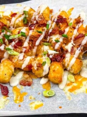 A hand picking up a cheese-covered tater tot from a tray. The tater tots are topped with melted cheese, sour cream drizzle, chopped green onions, and bacon bits, all arranged on a parchment-lined surface.