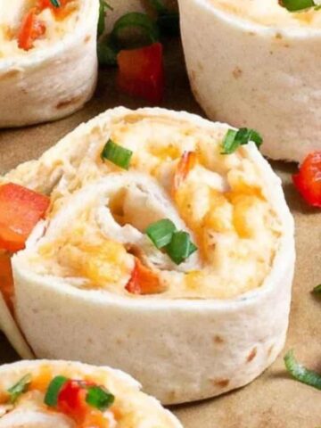 Tortilla pinwheels filled with cheese, red bell pepper, and green onions are arranged on a brown surface. A lemon wedge is placed alongside the pinwheels.