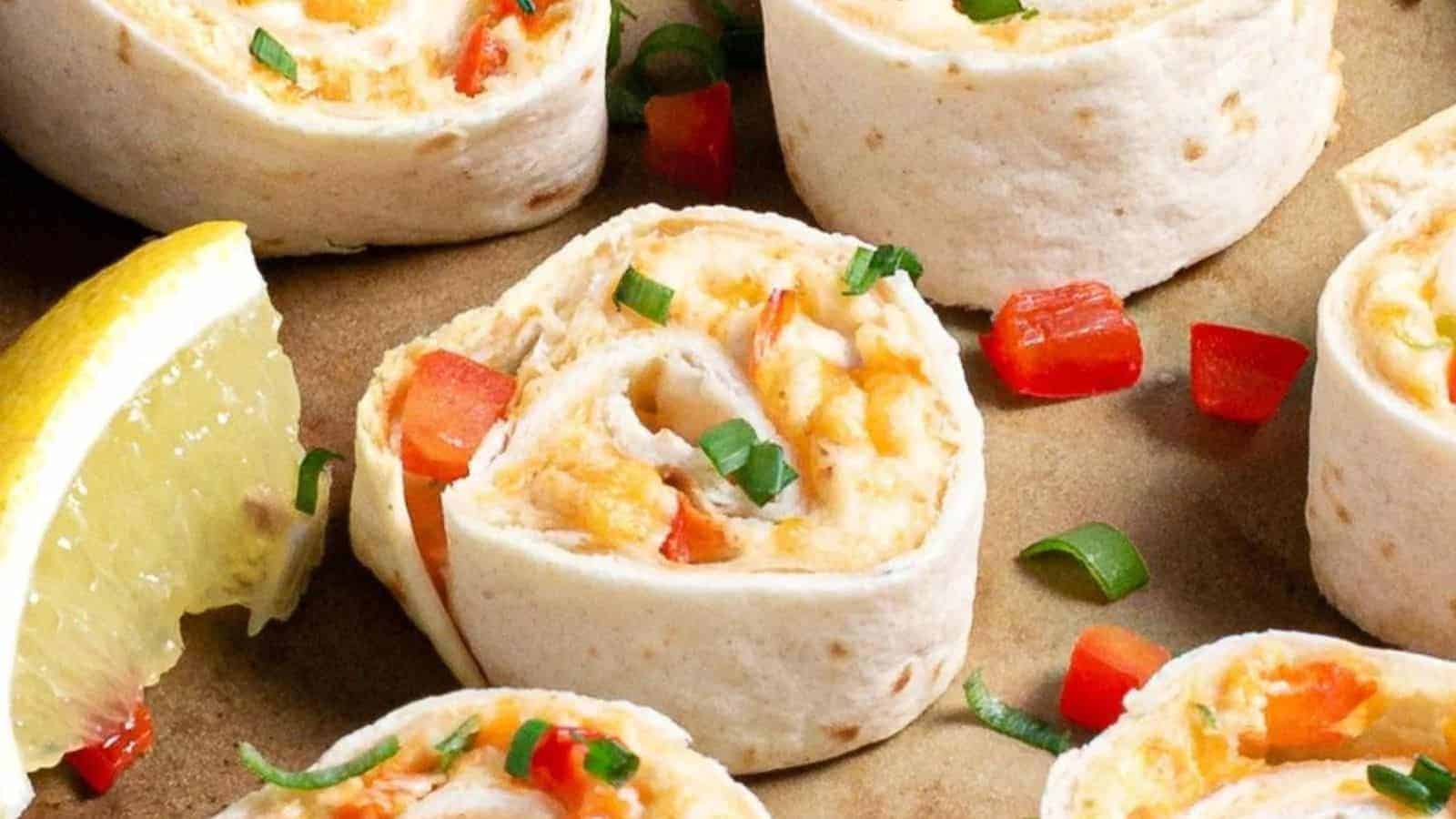 Tortilla pinwheels filled with cheese, red bell pepper, and green onions are arranged on a brown surface. A lemon wedge is placed alongside the pinwheels.