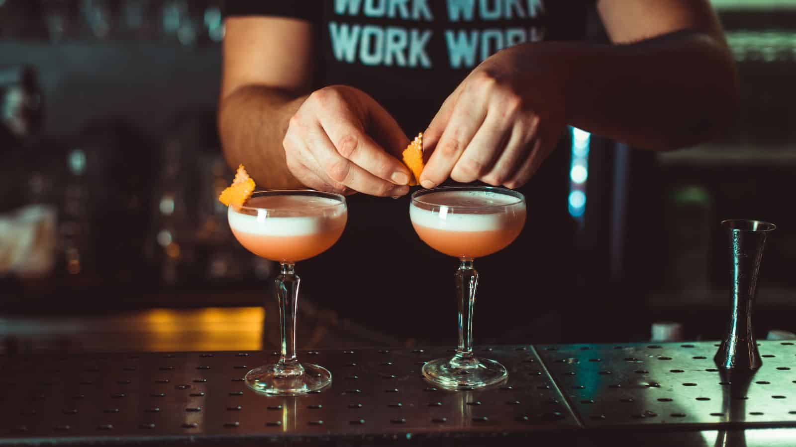 A bartender garnishes two cocktails on a bar counter. Each stemmed glass contains a pink drink with a frothy top, being adorned with an orange peel. The bartender wears a t-shirt with the word "WORK" repeated on it.