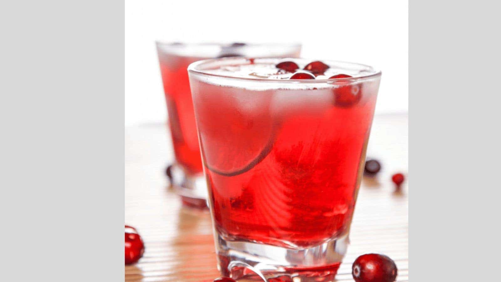 Two glasses filled with a red cranberry drink, garnished with whole cranberries, sit on a wooden surface. A few cranberries are scattered around the glasses. The background is softly blurred.