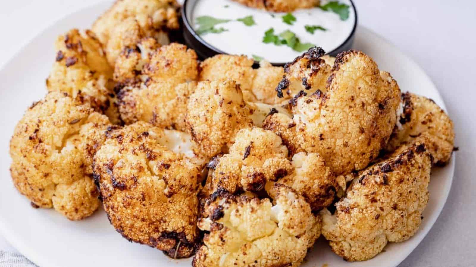 A plate of roasted cauliflower florets seasoned with spices, accompanied by a small bowl of creamy white sauce garnished with chopped parsley.